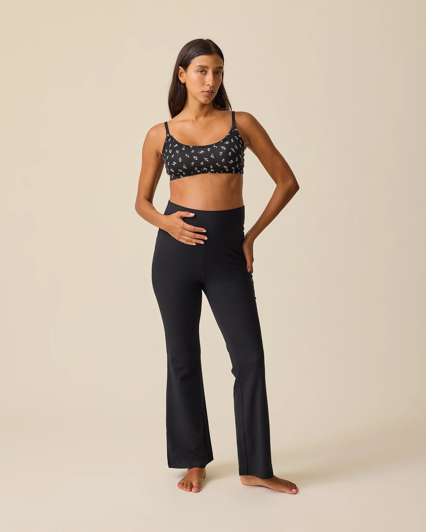 Front view of model wearing French Terry Racerback Nursing & Sleep Bra in Black Wildflower and Bamboo Flex Maternity & Postpartum Flare Pant in Black on a beige background.