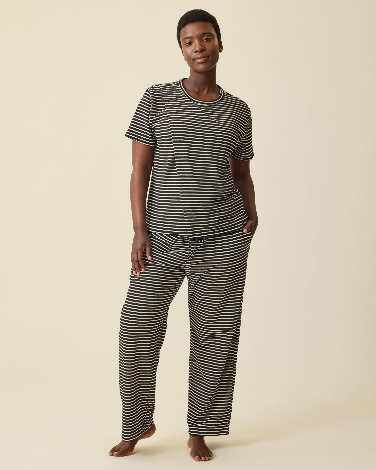 Model is wearing Luxe Terry Maternity & Nursing Split Hem T-shirt in Black & Ivory Stripe and Luxe Terry Maternity & Postpartum Pant in Black & Ivory Stripe, on a beige background.