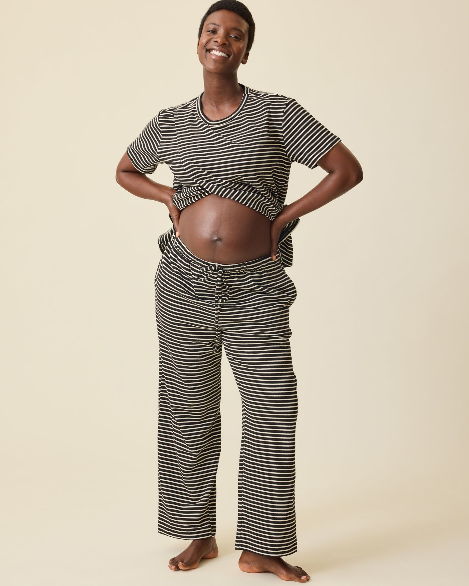 Model is showing baby bump and wearing Luxe Terry Maternity & Nursing Split Hem T-shirt in Black & Ivory Stripe and Luxe Terry Maternity & Postpartum Pant in Black & Ivory Stripe, on a beige background.