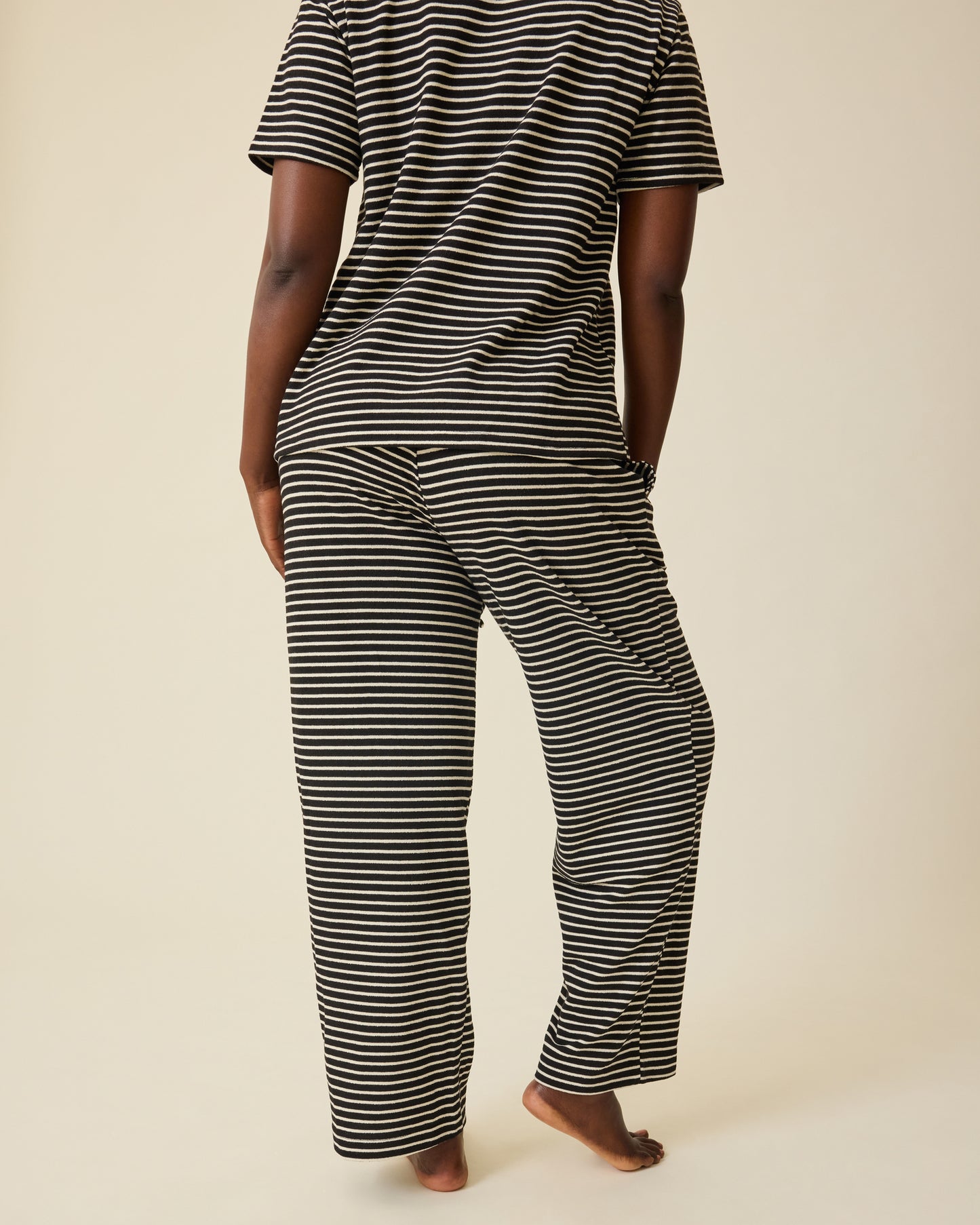 Back view of model wearing Luxe Terry Maternity & Nursing Split Hem T-shirt in Black & Ivory Stripe and Luxe Terry Maternity & Postpartum Pant in Black & Ivory Stripe, on a beige background.