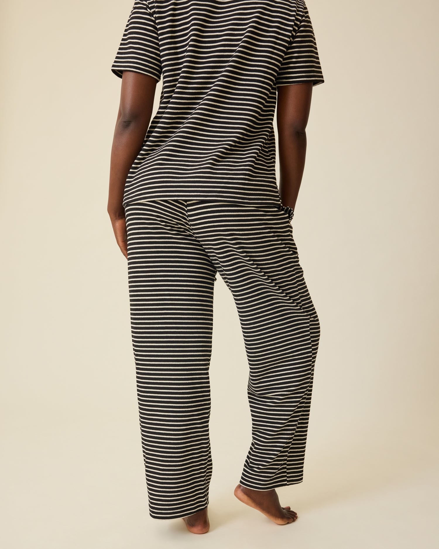 Back view of model wearing Luxe Terry Maternity & Nursing Split Hem T-shirt in Black & Ivory Stripe and Luxe Terry Maternity & Postpartum Pant in Black & Ivory Stripe, on a beige background.