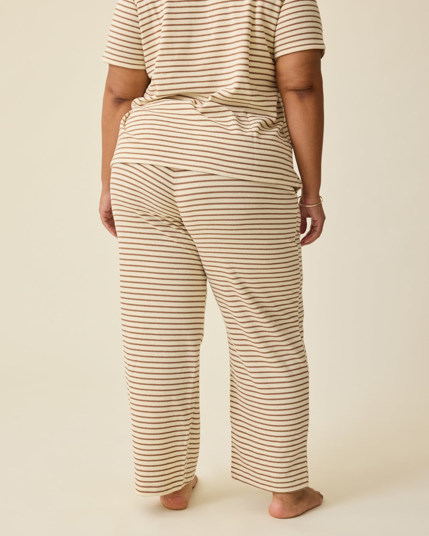 Back view of Model wearing Luxe Terry Maternity & Nursing Split Hem T-shirt in Ivory & Chocolate Stripe and Luxe Terry Maternity & Postpartum Pant in Ivory & Chocolate Stripe, on a beige background.