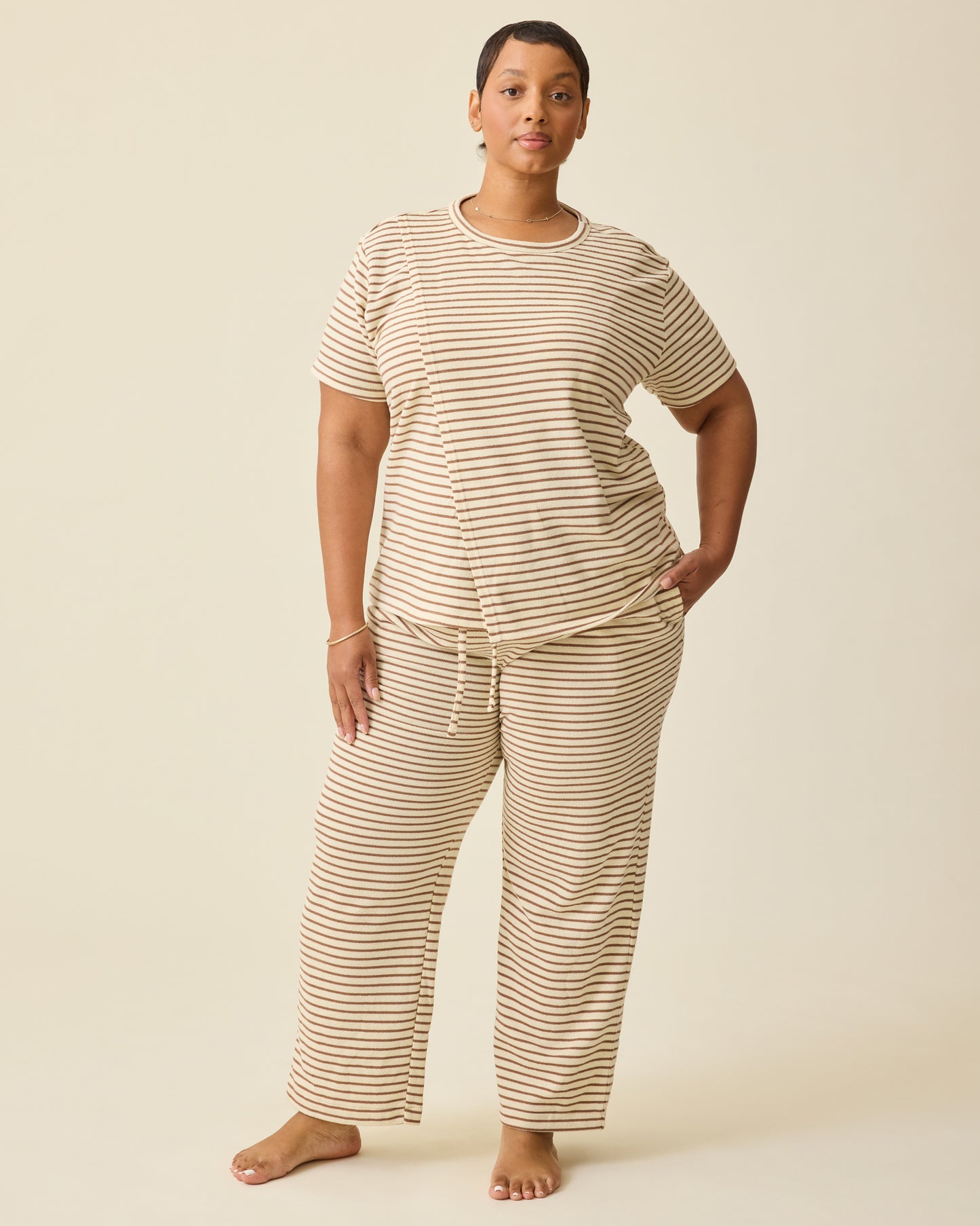 Model is wearing Luxe Terry Maternity & Nursing Split Hem T-shirt in Ivory & Chocolate Stripe and Luxe Terry Maternity & Postpartum Pant in Ivory & Chocolate Stripe, on a beige background. 