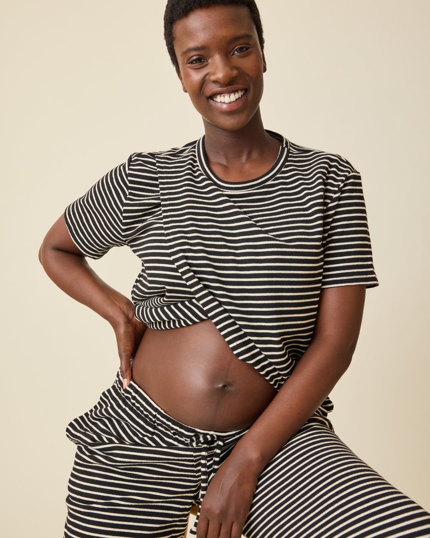 Model is showing baby bump and wearing Luxe Terry Maternity & Nursing Split Hem T-shirt in Black & Ivory Stripe and Luxe Terry Maternity & Postpartum Pant in Black & Ivory Stripe, on a beige background.