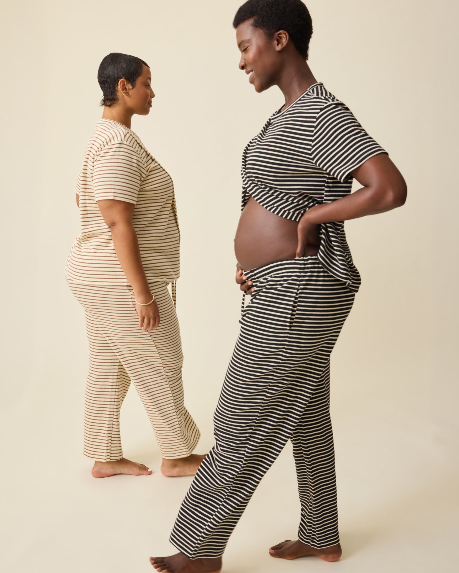 Model on right is wearing Luxe Terry Maternity & Nursing Split Hem T-shirt in Black & Ivory Stripe and Luxe Terry Maternity & Postpartum Pant in Black & Ivory Stripe, and model on left is wearing  Luxe Terry Maternity & Nursing Split Hem T-shirt in Ivory & Chocolate Stripe and Luxe Terry Maternity & Postpartum Pant in Ivory & Chocolate Stripe, on a beige background.