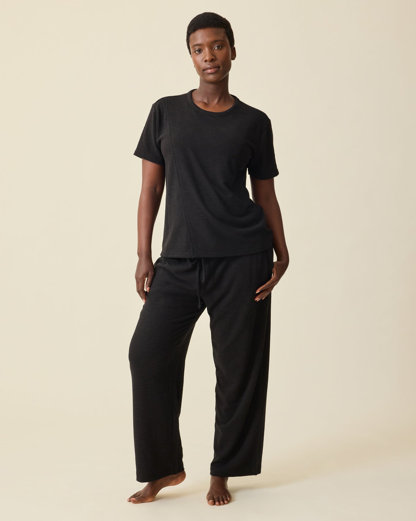 Model is wearing Luxe Terry Maternity & Nursing Split Hem T-shirt in Black and Luxe Terry Maternity & Postpartum Pant in Black, on a beige background.