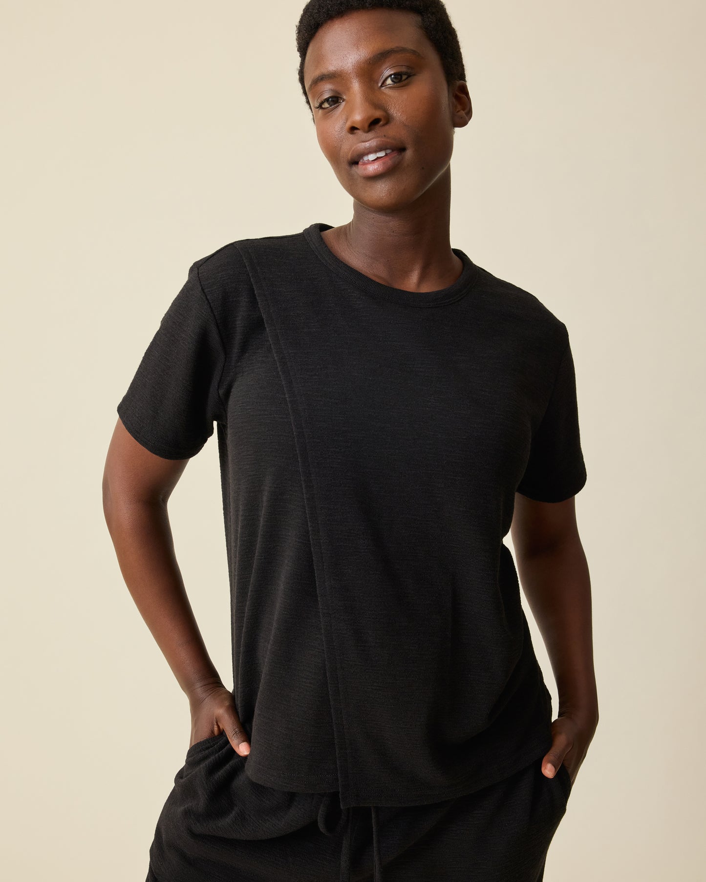 Model is wearing Luxe Terry Maternity & Nursing Split Hem T-shirt in Black and Luxe Terry Maternity & Postpartum Pant in Black, on a beige background.@model_info:Anna is wearing a Small.