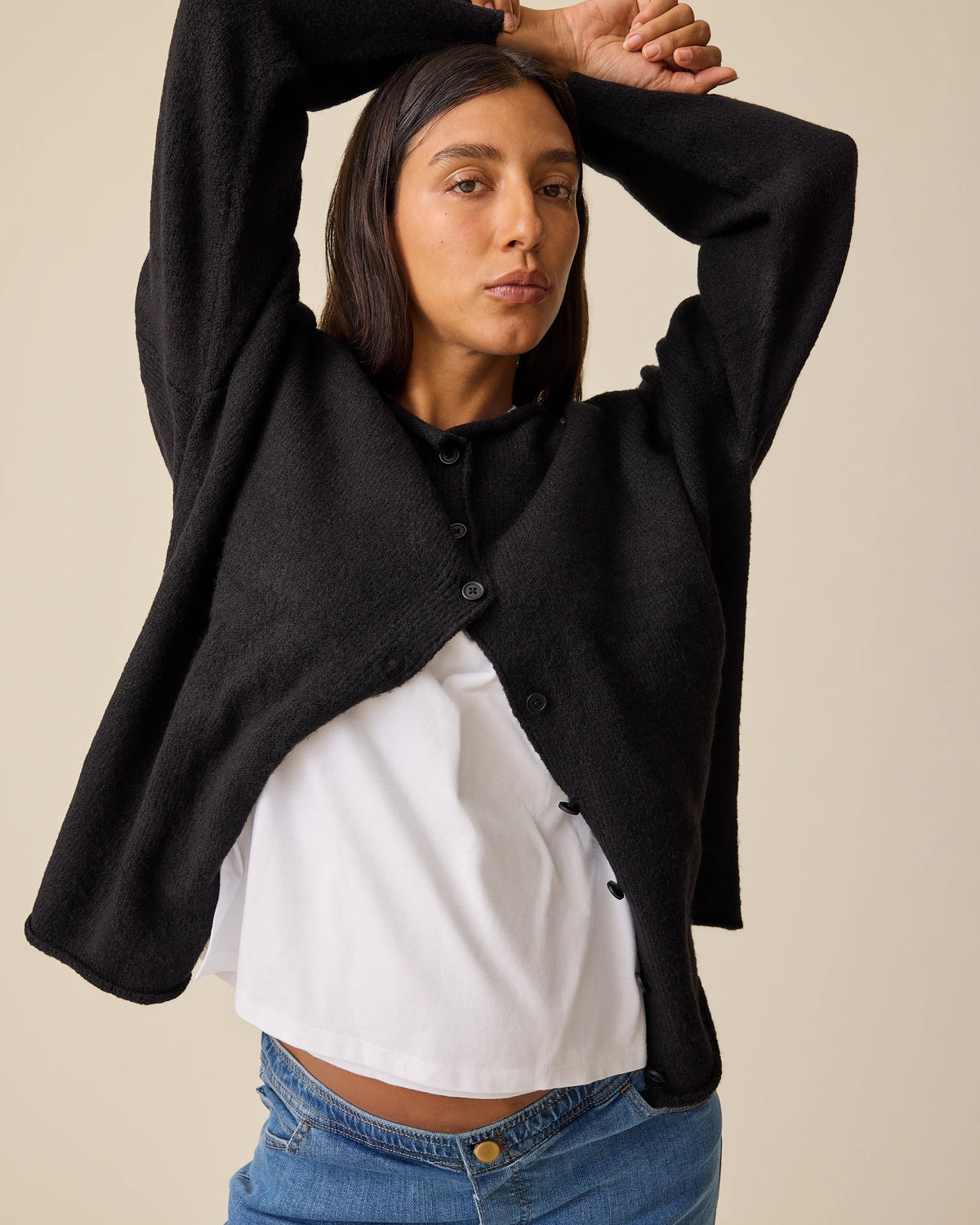 Model is holding arms over head, showing a peek of baby bump while wearing Ultra Soft Maternity & Postpartum Cardigan in Black, Wide Leg Maternity & Postpartum Jean in Medium Wash and a white nursing top, on a beige background.