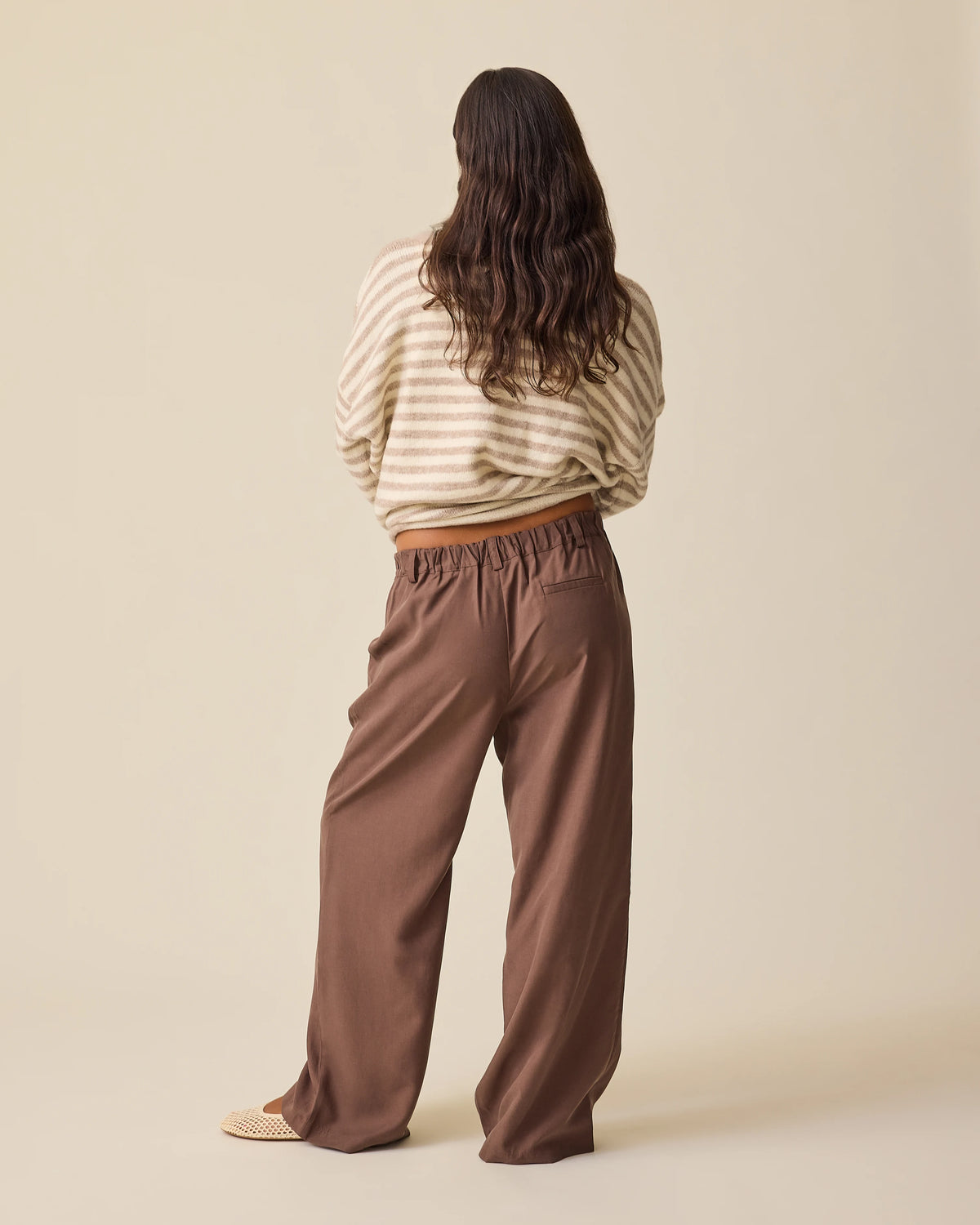 Back view of model wearing Ultra Soft Maternity & Postpartum Cardigan in Tan & White Stripe, Maternity & Postpartum Wide Leg Trouser in Brown and a nursing white tee, on a beige background