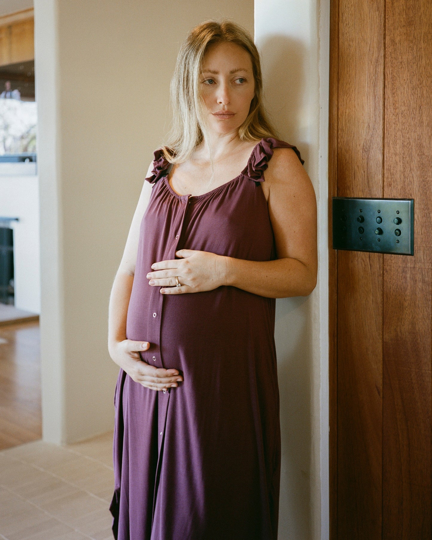 Pregnant woman wearing the Ruffle Strap Labor & Delivery Gown in Burgundy Plum, standing in a light filled midcentury home.@model_info:Kate is 5'7" and wearing a Small.