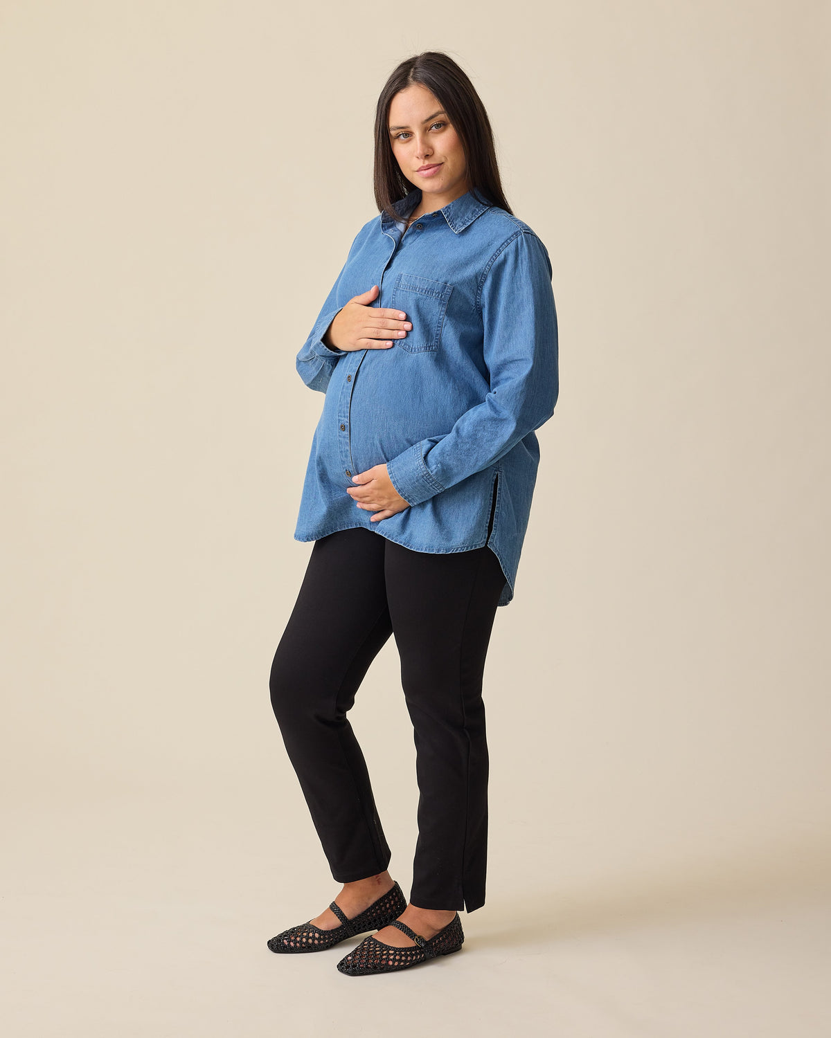 Model is wearing Denim Maternity & Nursing Button Up Shirt in Medium Wash and Maternity Ponte Straight Leg Pant in Black on a beige background.