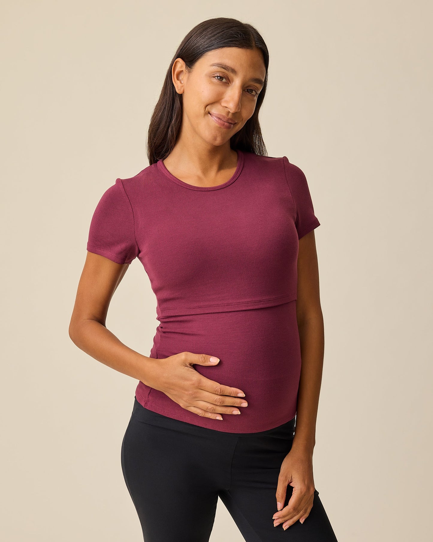 Model is wearing Ribbed Bamboo Crew Neck Nursing T-shirt in Deep Burgundy with black pants on a beige background. @model_info:Hayley is wearing a Small.