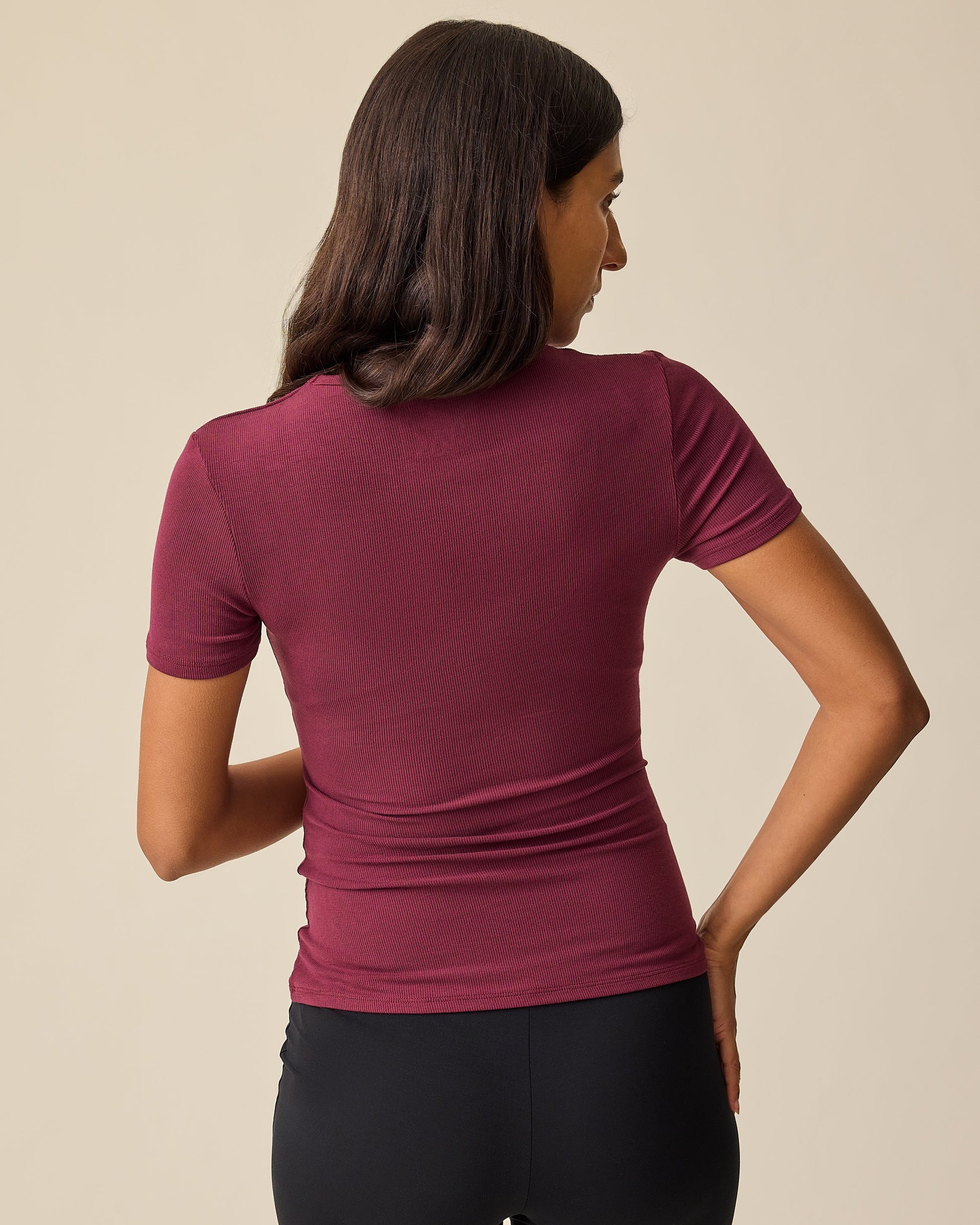 Back view of model wearing Ribbed Bamboo Crew Neck Nursing T-shirt in Deep Burgundy with black pants on a beige background.