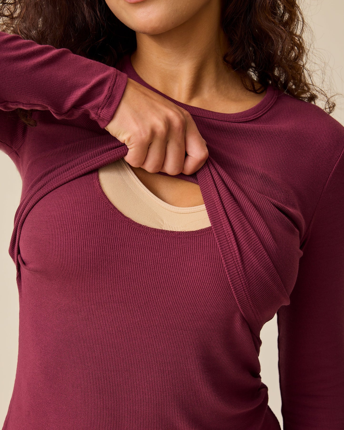 Model is lifting panel for easy nursing access on the Ribbed Bamboo Long Sleeve Nursing T-shirt in Deep Burgundy with black pants, on a beige background.