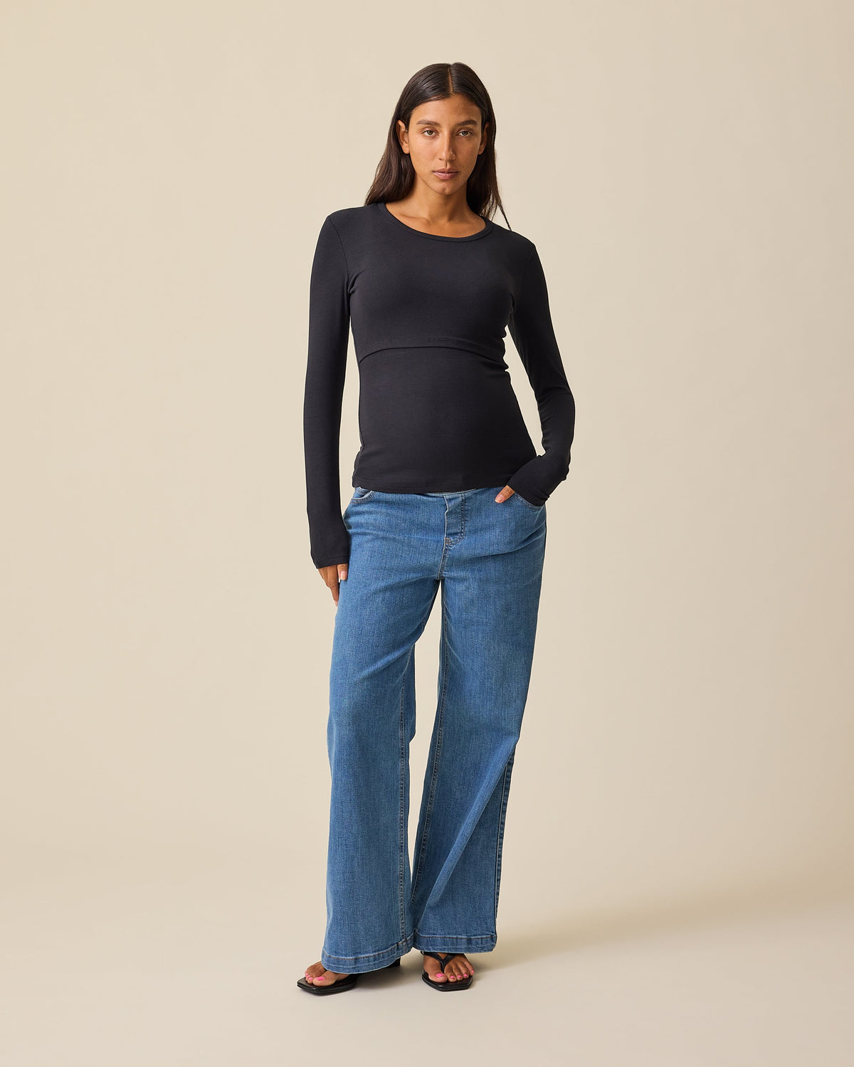 A woman wearing the Ribbed Bamboo Long Sleeve Nursing T-shirt in Black,  paired with blue jeans, against a solid beige background 