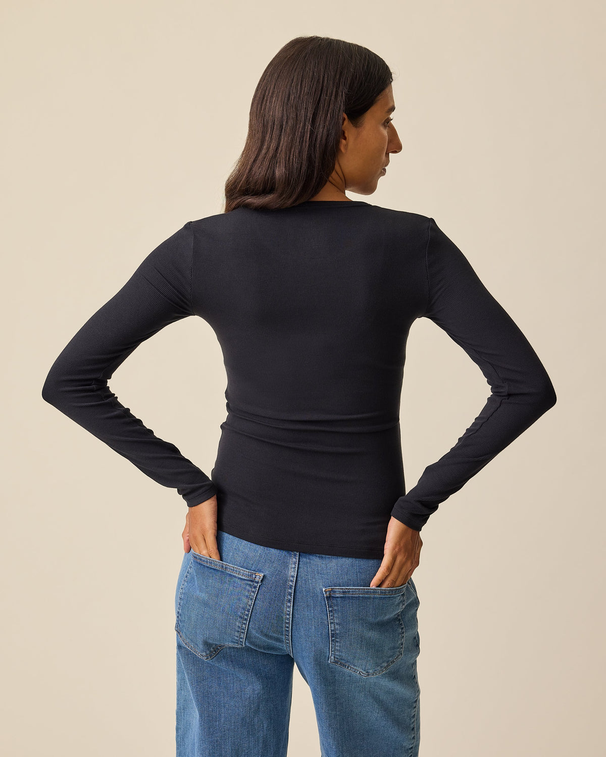 A woman showing the back of the Ribbed Bamboo Long Sleeve Nursing T-shirt in Black,  paired with blue jeans, against a solid beige background 