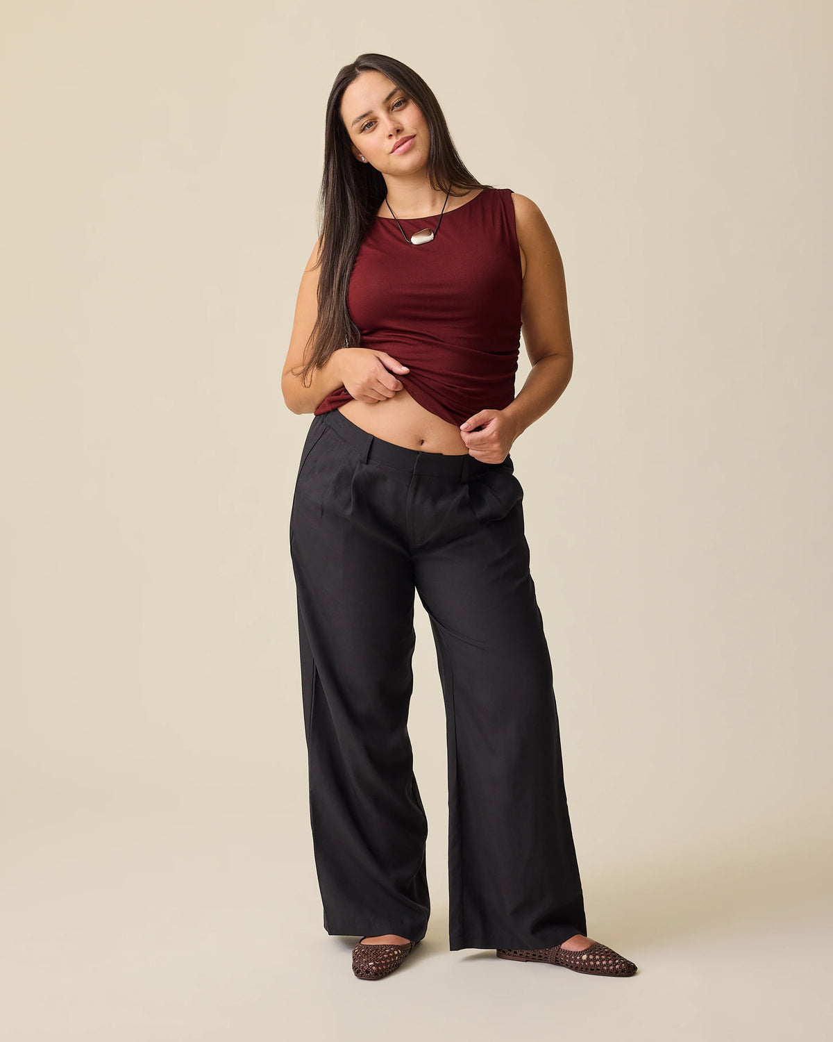 Model is wearing Ruched Maternity Sleeveless Top in Burgundy and Maternity & Postpartum Wide Leg Trouser in Black on a beige background.@model_info:Brianna is 5'8" and wearing a Medium.