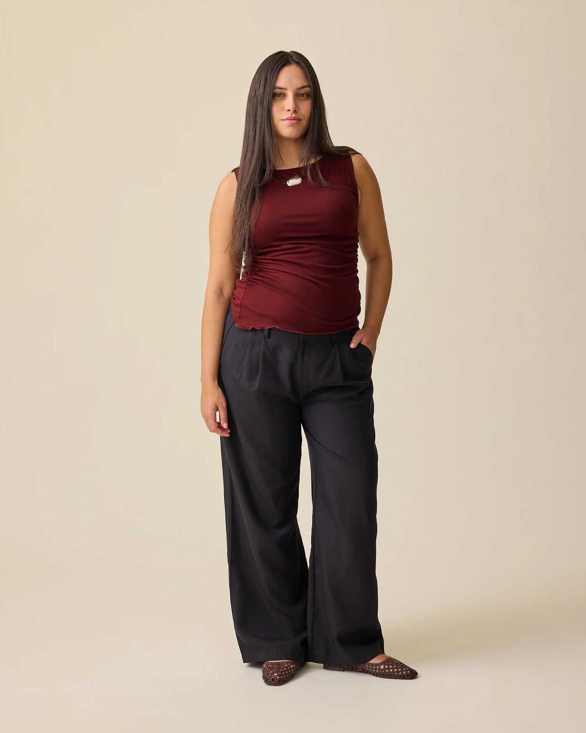 Model is wearing Ruched Maternity Sleeveless Top in Burgundy and Maternity & Postpartum Wide Leg Trouser in Black on a beige background. 
