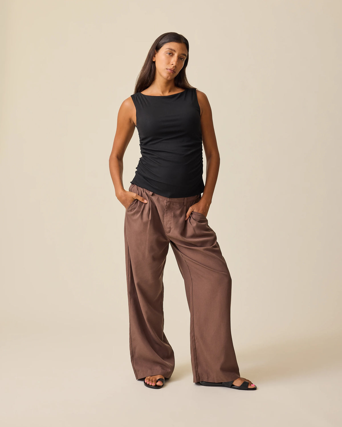 Model is wearing Maternity & Postpartum Wide Leg Trouser in Brown and Ruched Maternity Sleeveless Top in Black, on a beige background.