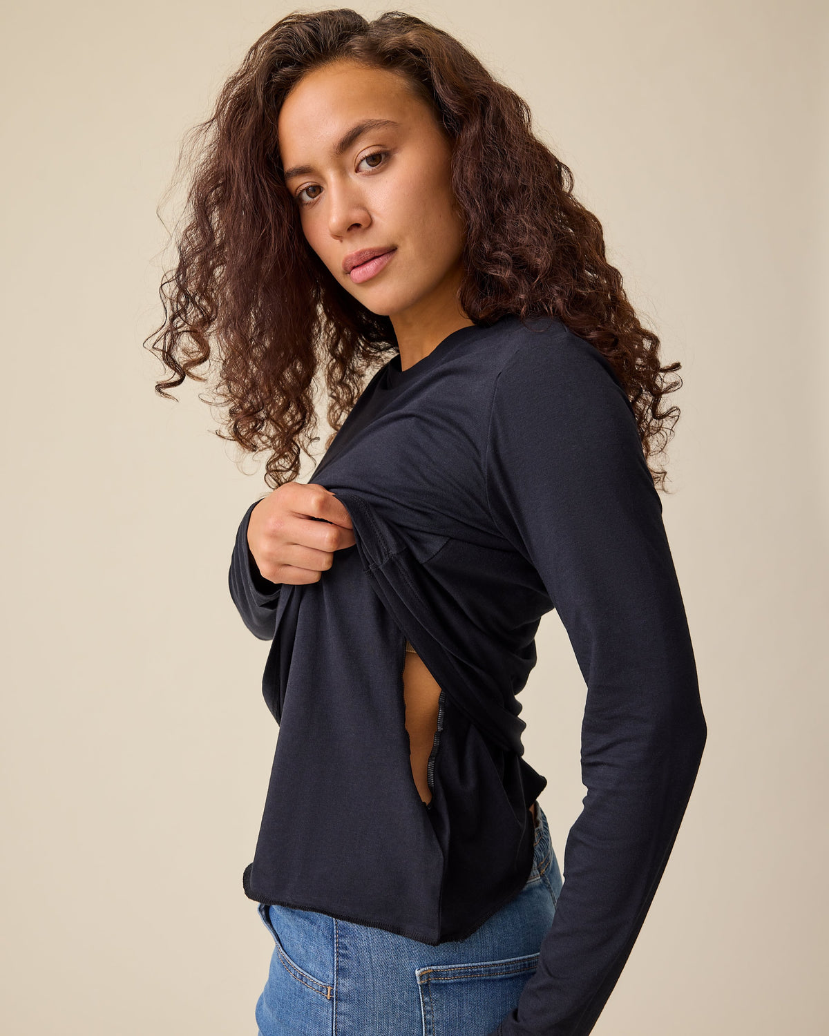 Side view of model lifting front panel to display easy nursing access Modal Long Sleeve Nursing T-shirt in Black and Wide Leg Maternity & Postpartum Jean in Medium Wash on a beige background.