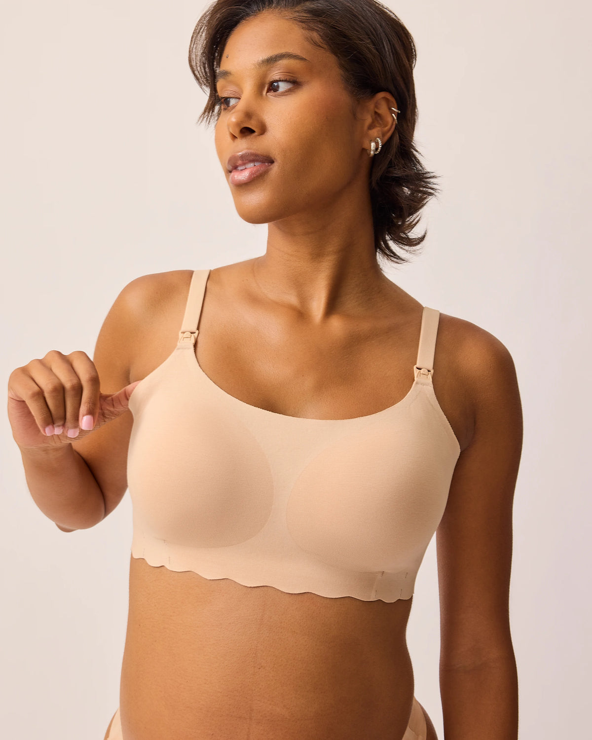 Model is wearing BareBond™ Cotton Maternity & Nursing Bralette in Sand while stretching the side to show soft material, on a beige background.@model_info:Ale is wearing a Medium.