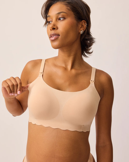 Model is wearing BareBond™ Cotton Maternity & Nursing Bralette in Sand while stretching the side to show soft material, on a beige background.@model_info:Ale is wearing a Medium.