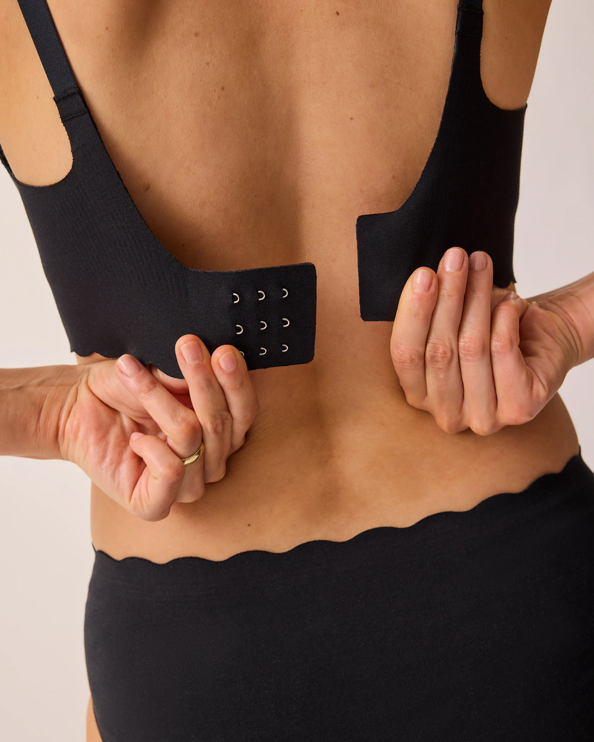 Back view of model putting on the BareBond™ Cotton Maternity & Nursing Plunge Bra in Black on a beige background.