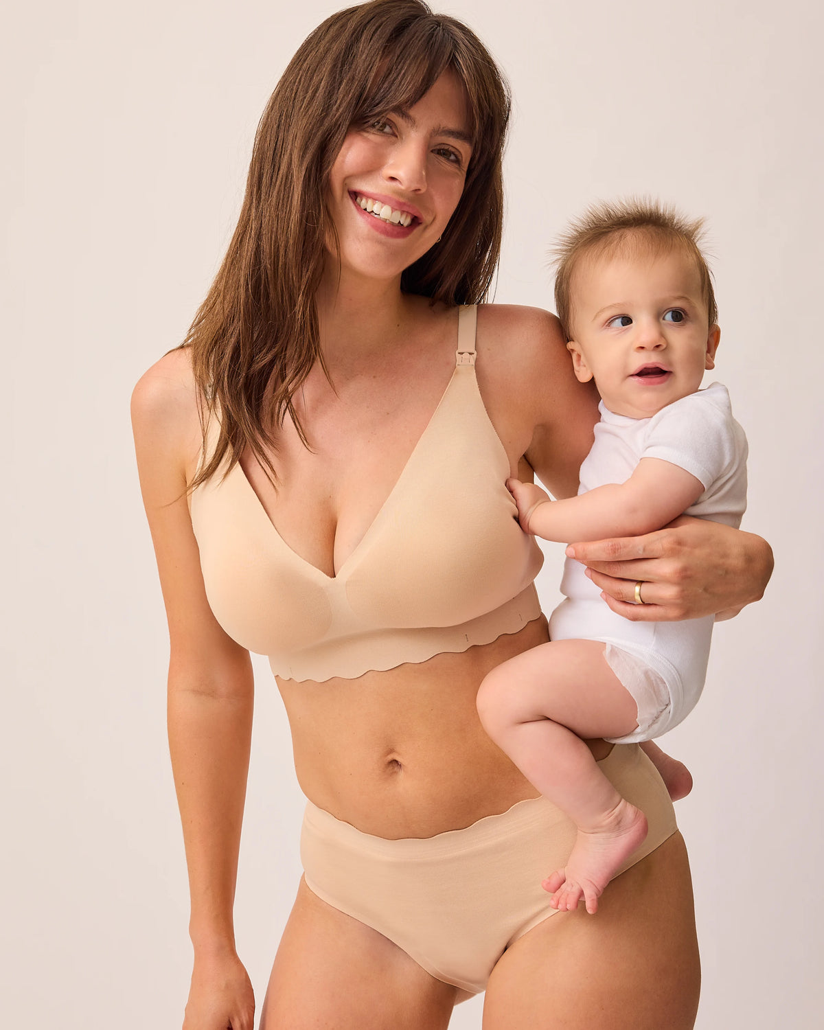 Model is holding baby and wearing BareBond™ Cotton Maternity & Nursing Plunge Bra in Sand and BareBond™ Cotton Maternity & Postpartum Hipster in Sand on a beige background.