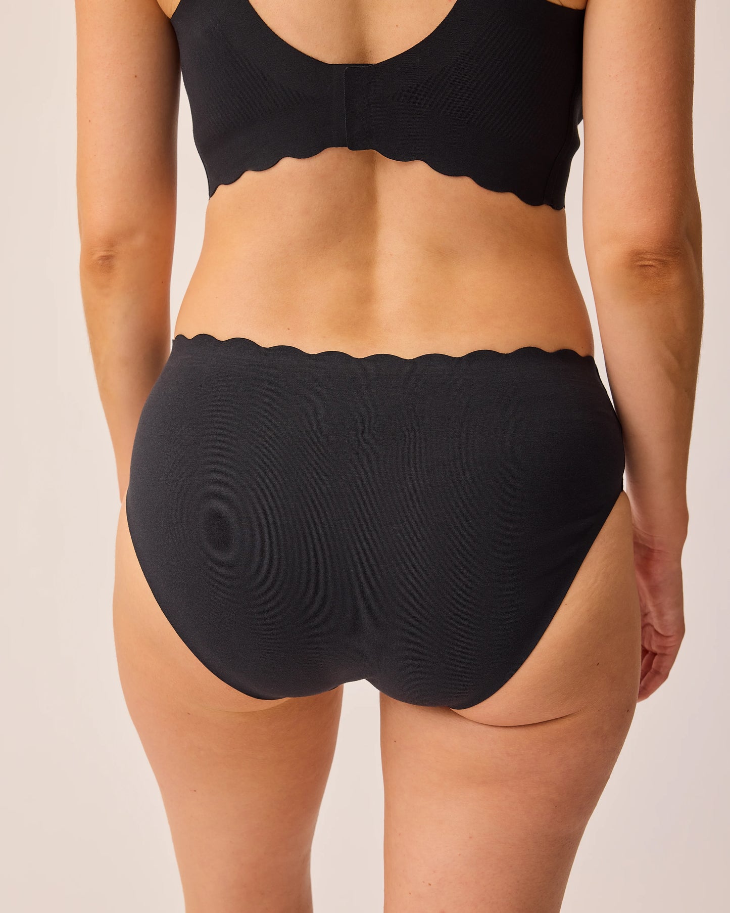 Back view of model wearing BareBond™ Cotton Maternity & Postpartum Hipster in Black and BareBond™ Cotton Maternity & Nursing Bra in Black on a beige background.
