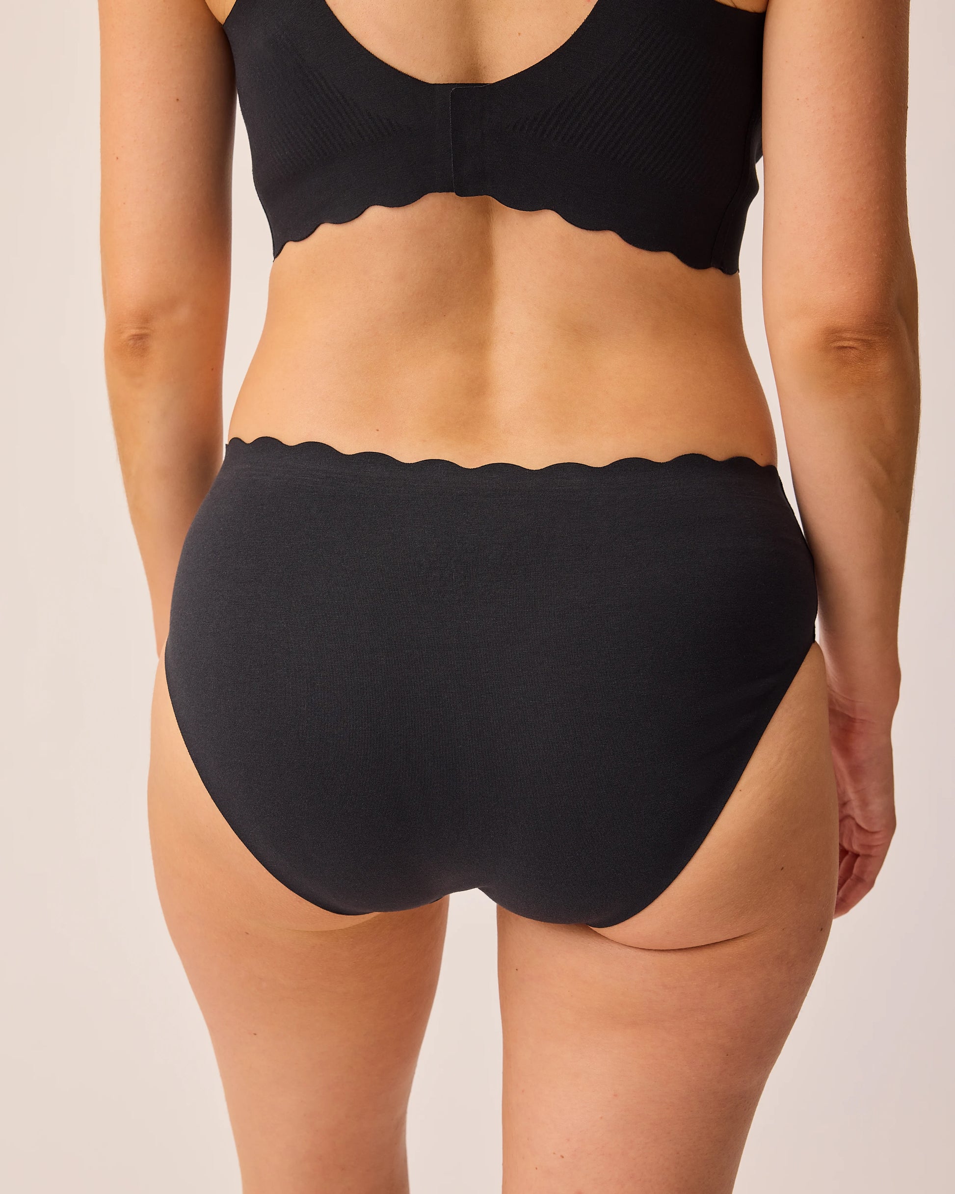 Back view of model wearing BareBond™ Cotton Maternity & Postpartum Hipster in Black and BareBond™ Cotton Maternity & Nursing Bra in Black on a beige background.