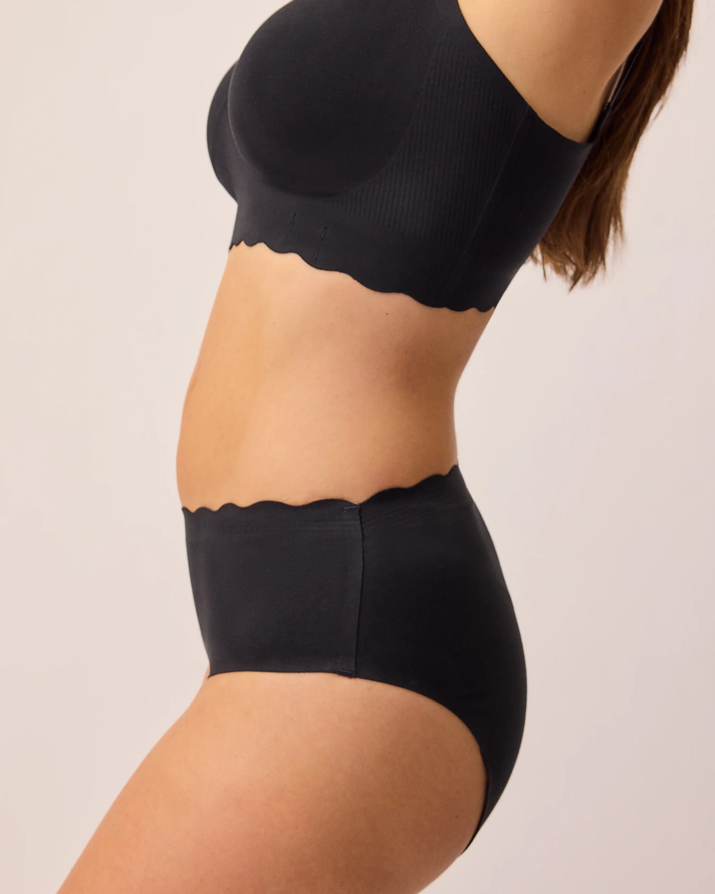Side view of model wearing BareBond™ Cotton Maternity & Postpartum Brief in Black and BareBond™ Cotton Maternity & Nursing Bra in Black on a beige background.