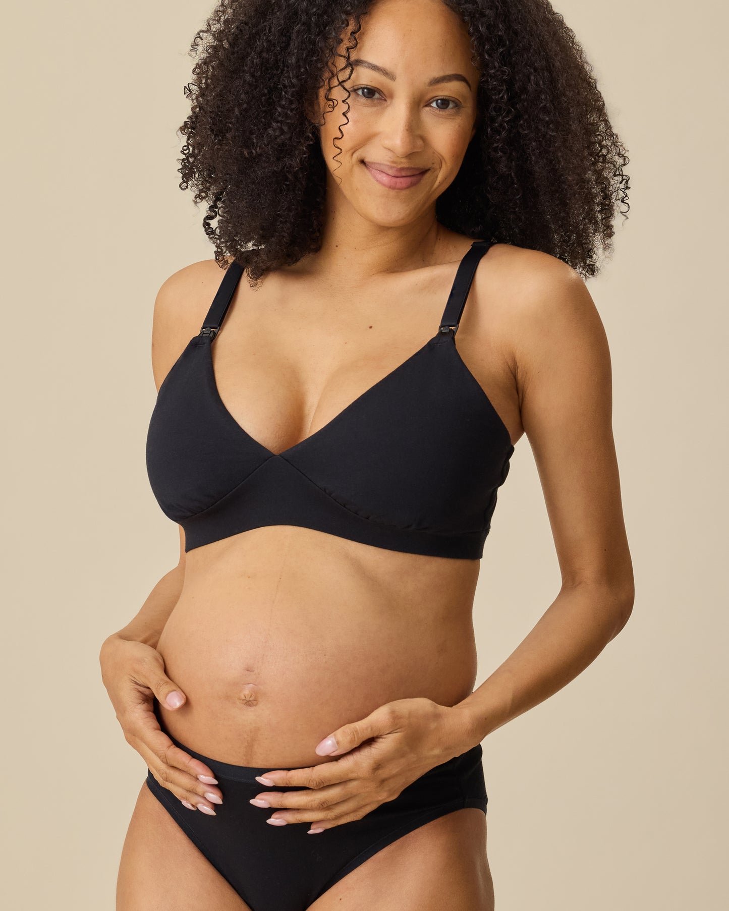 Model is wearing SoftStretch Cotton Maternity & Nursing Bralette in Black and SoftStretch Cotton Maternity & Postpartum Brief in Black on a beige background.