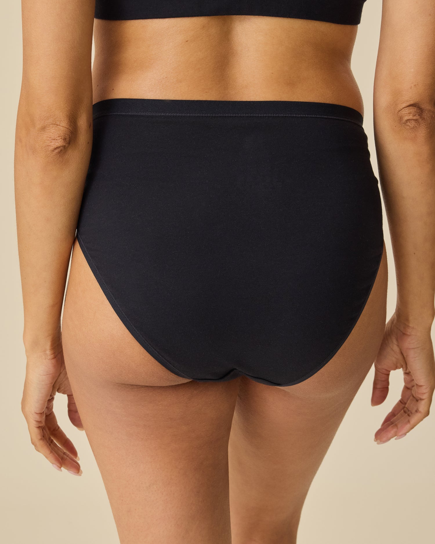 Back view of model wearing SoftStretch Cotton Maternity & Nursing Bralette in Black and SoftStretch Cotton Maternity & Postpartum Brief in Black on a beige background.