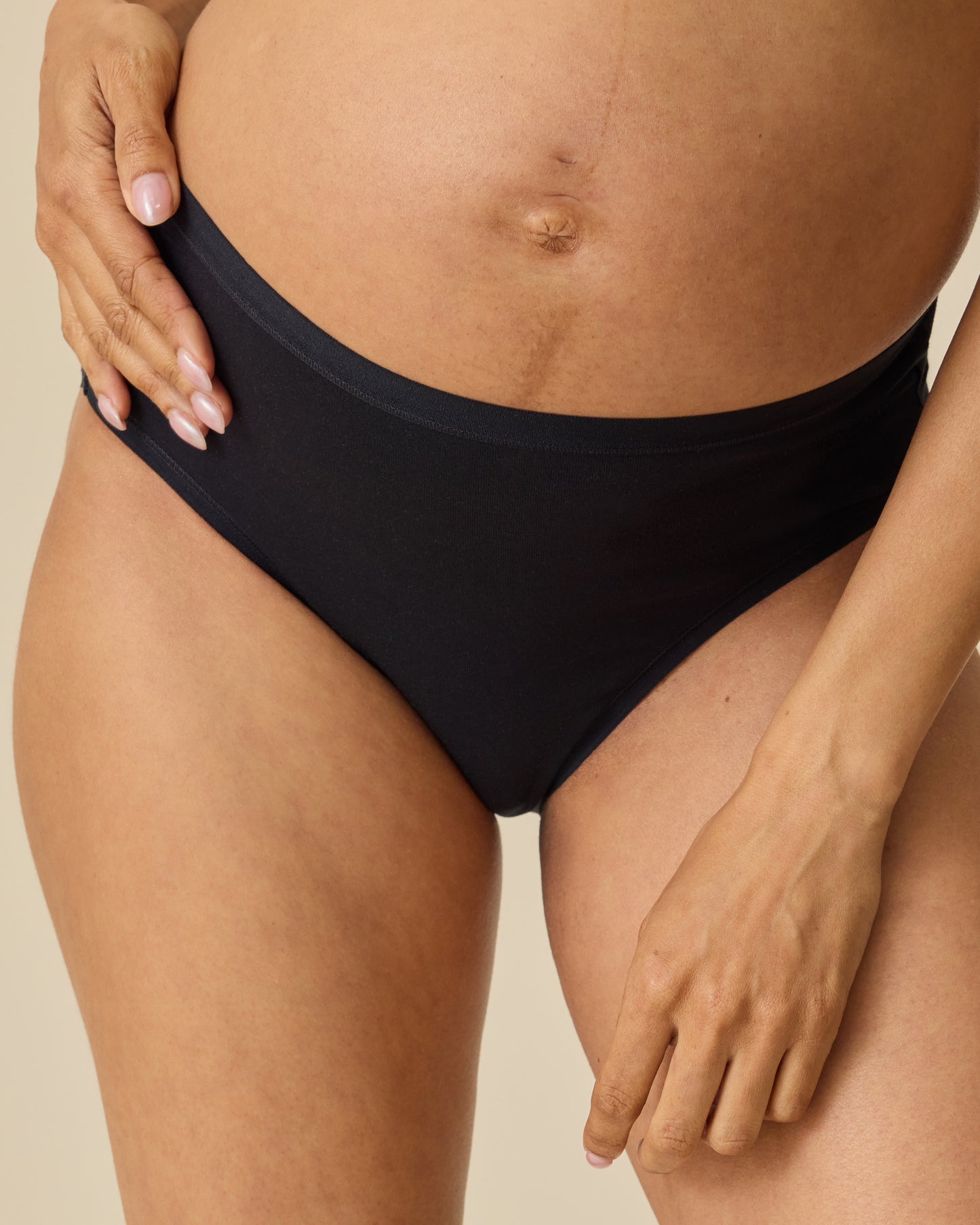 Close up of model wearing SoftStretch Cotton Maternity & Postpartum Brief in Black on a beige background.@model_info:Courtney is wearing a Small.