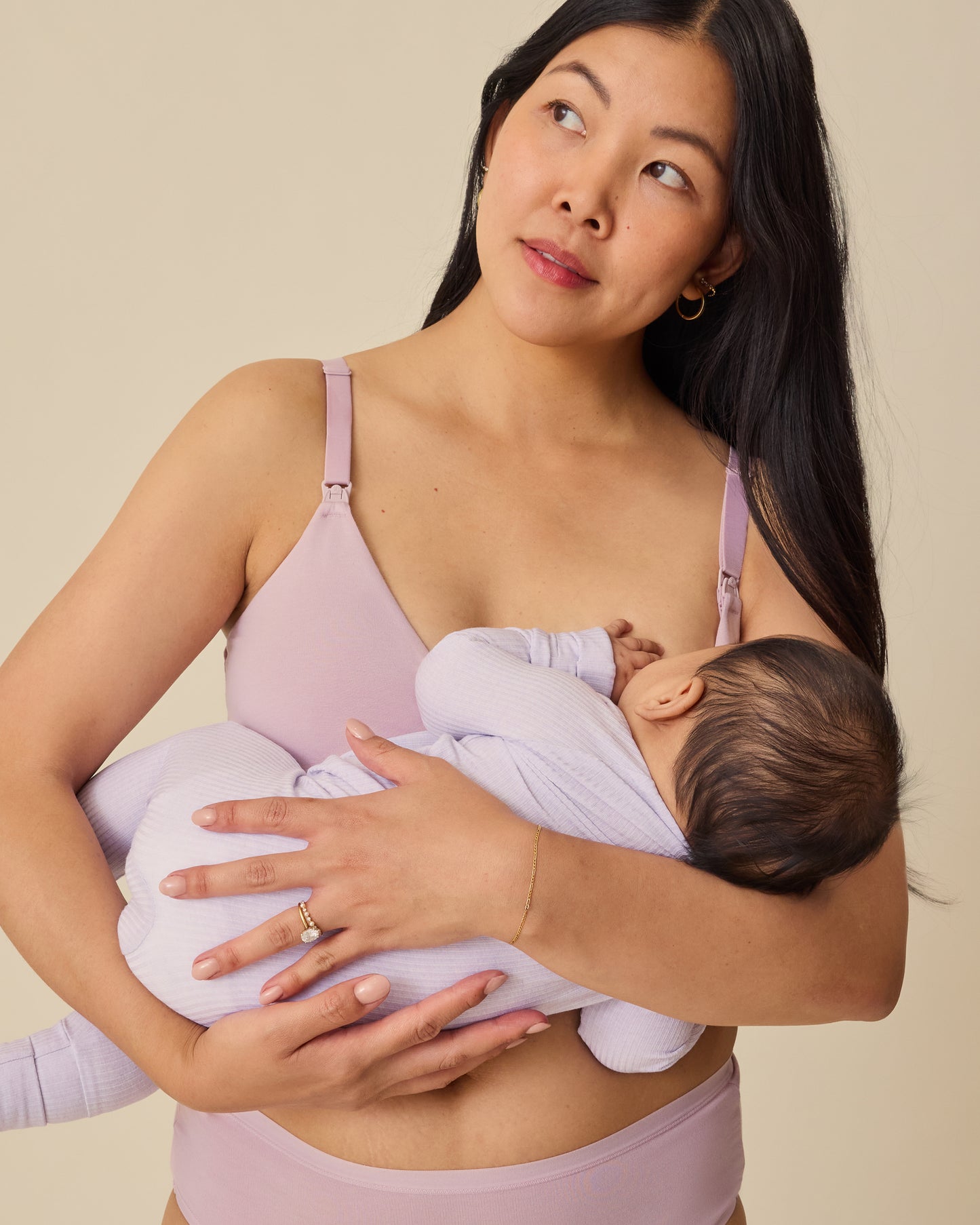 Model is breastfeeding baby in Ribbed Bamboo Baby Pajama in Lilac Heather while wearing SoftStretch Cotton Maternity & Nursing Bralette in Lilac Mist on a beige background. 