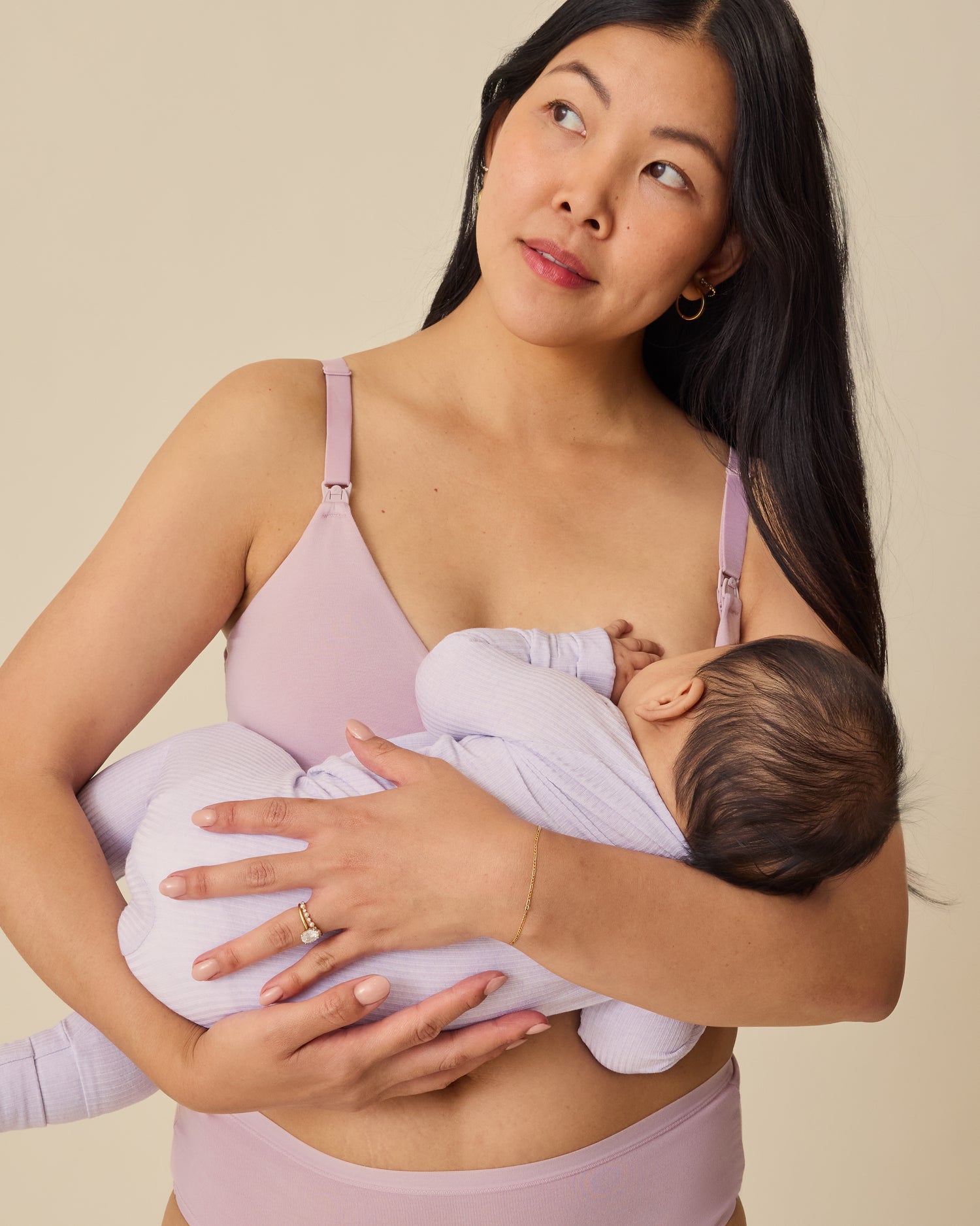 Model is breastfeeding baby in Ribbed Bamboo Baby Pajama in Lilac Heather while wearing SoftStretch Cotton Maternity & Nursing Bralette in Lilac Mist on a beige background. 