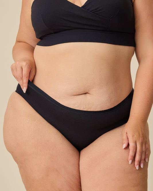 Model is wearing SoftStretch Cotton Nursing Bra in Black and SoftStretch Cotton Maternity & Postpartum Hipster in Black on a beige background.@model_info:Allie is wearing a 1X.
