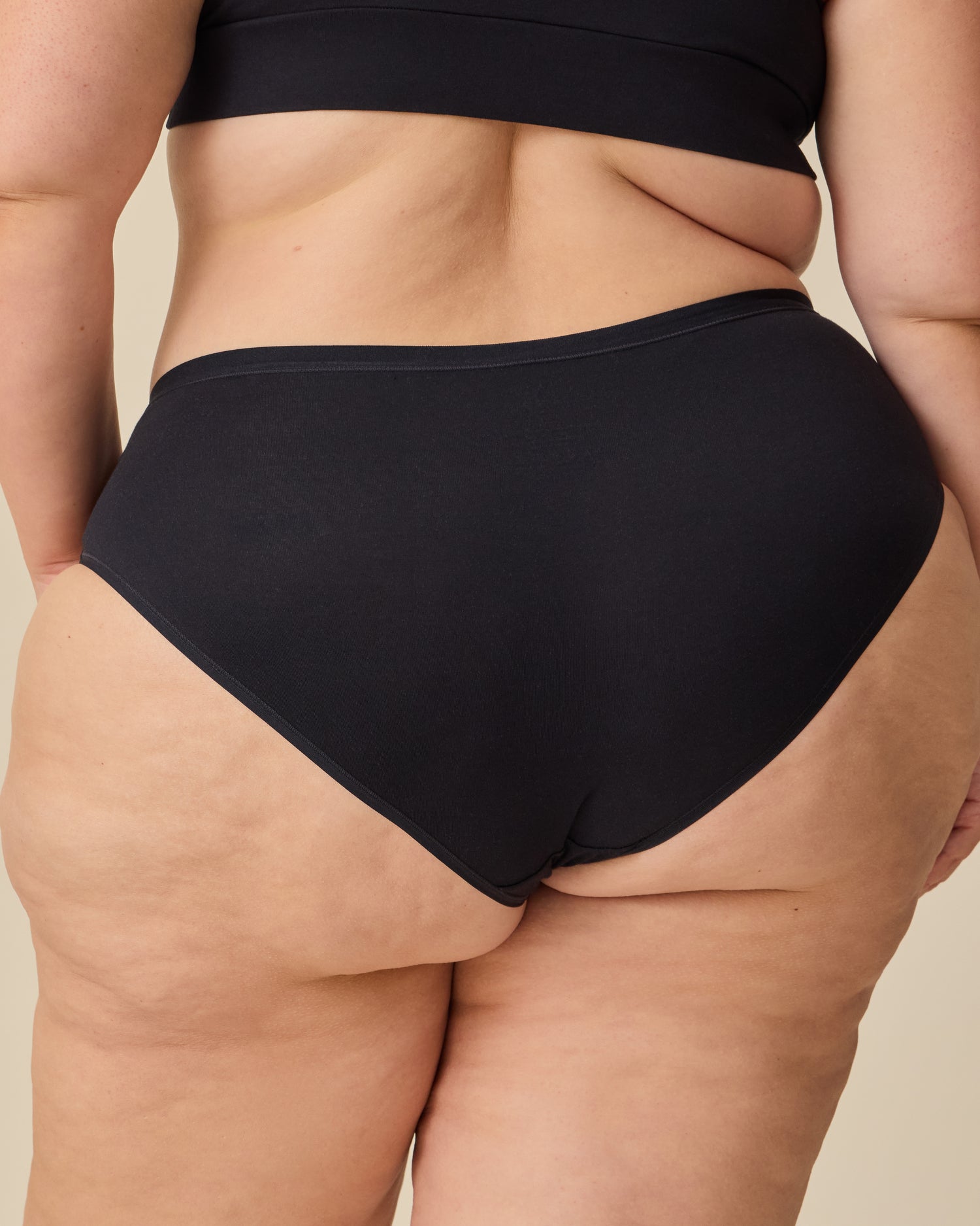 Back view of the model wearing SoftStretch Cotton Nursing Bra in Black and SoftStretch Cotton Maternity & Postpartum Hipster in Black on a beige background.