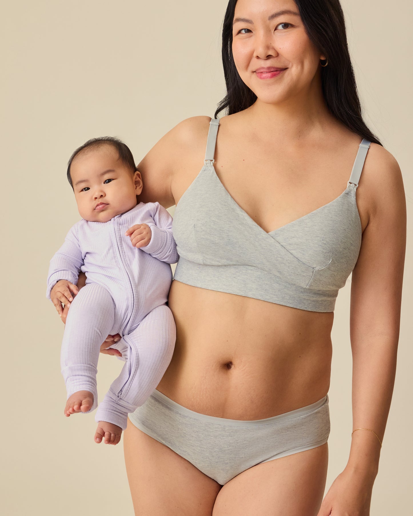 Model is holding her baby and wearing SoftStretch Cotton Nursing Bra in Grey Heather and SoftStretch Cotton Maternity & Postpartum Hipster in Grey Heather on a beige background. 