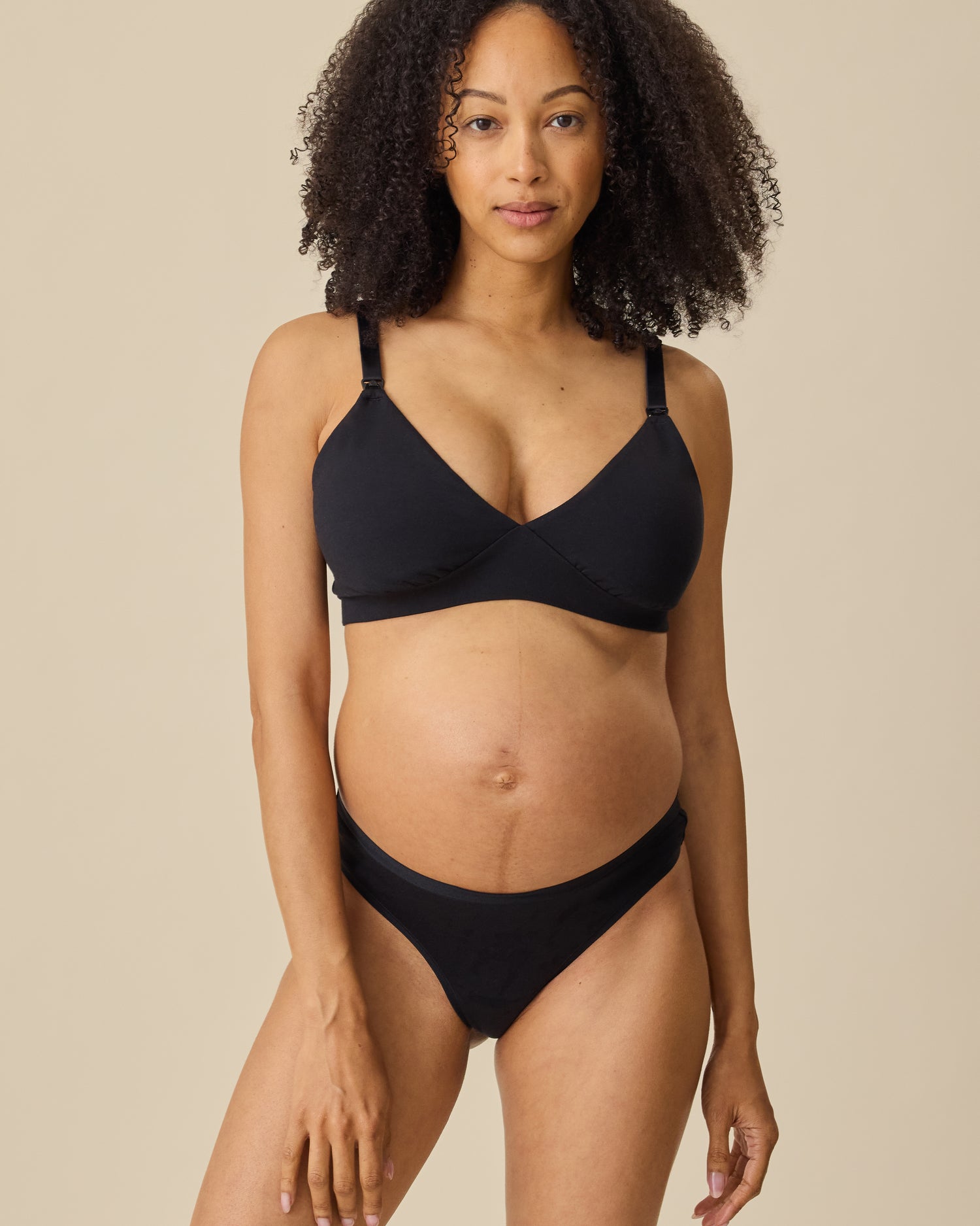 Model wearing the SoftStretch Cotton Maternity & Nursing Bralette in Black and SoftStretch Cotton Maternity & Postpartum Thong in Black, on a beige background.
