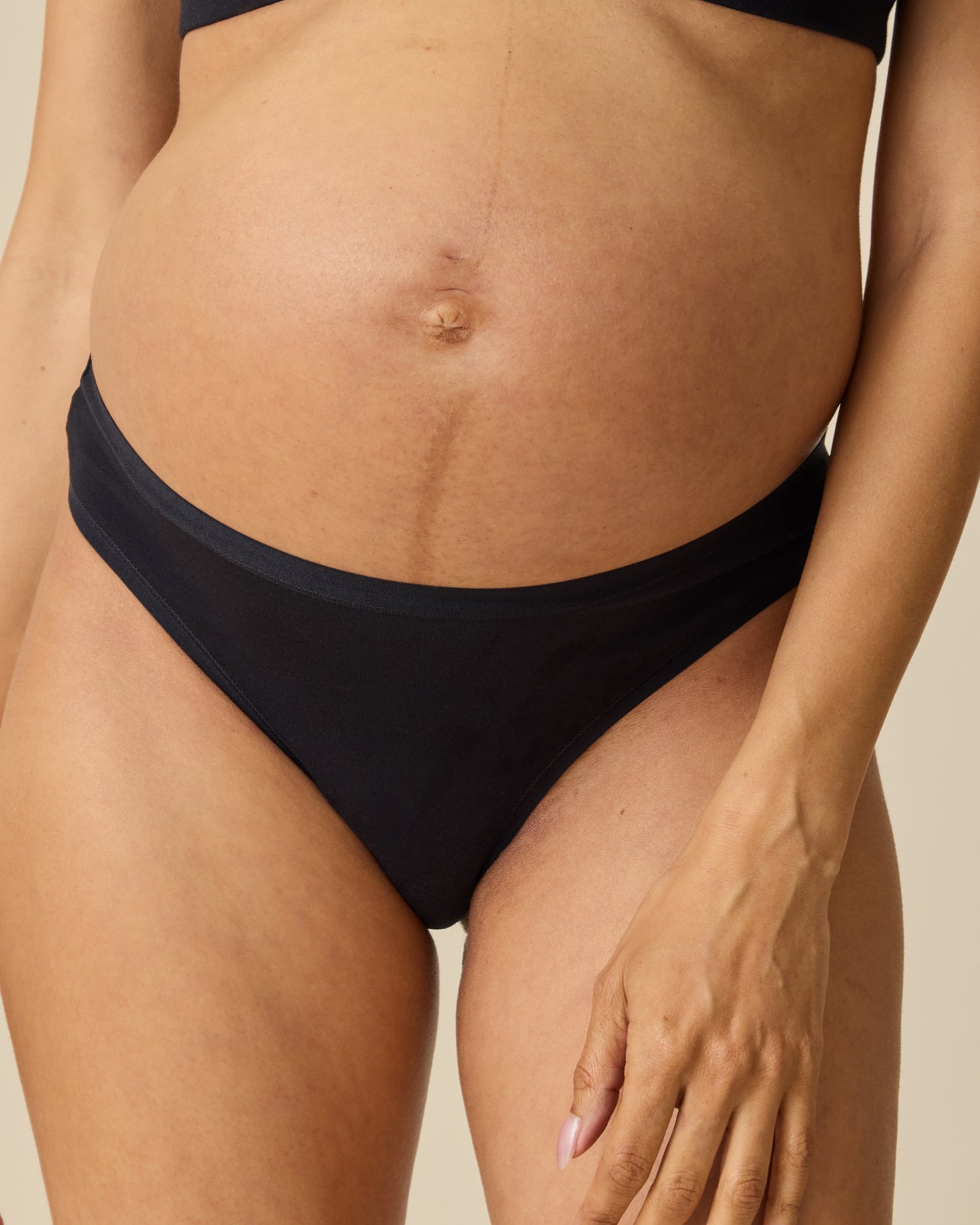 Model wearing the SoftStretch Cotton Maternity & Postpartum Thong in Black, on a beige background. @model_info:Courtney is wearing a Small.