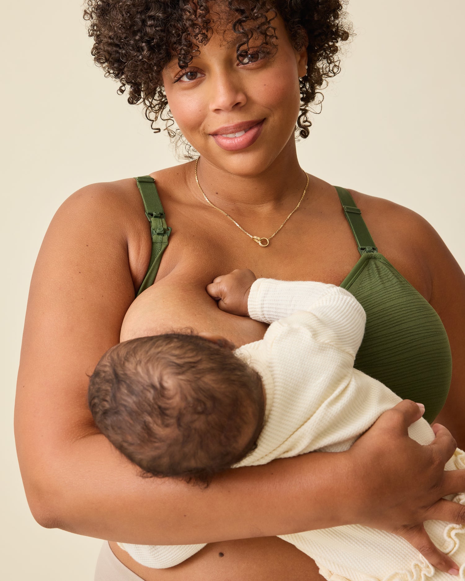 Model is breastfeeding while wearing Sublime® Hands-Free Pumping & Nursing Bra in Deep Olive on a beige background.