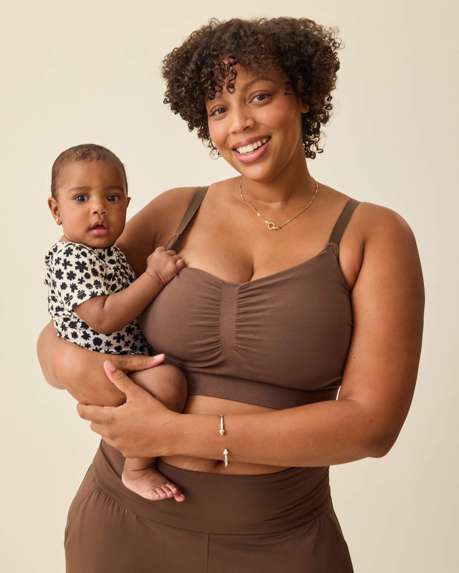 Model is holding baby while wearing Sublime® Bamboo Hands-Free Pumping Lounge & Sleep Bra in Chocolate on a beige background. 
