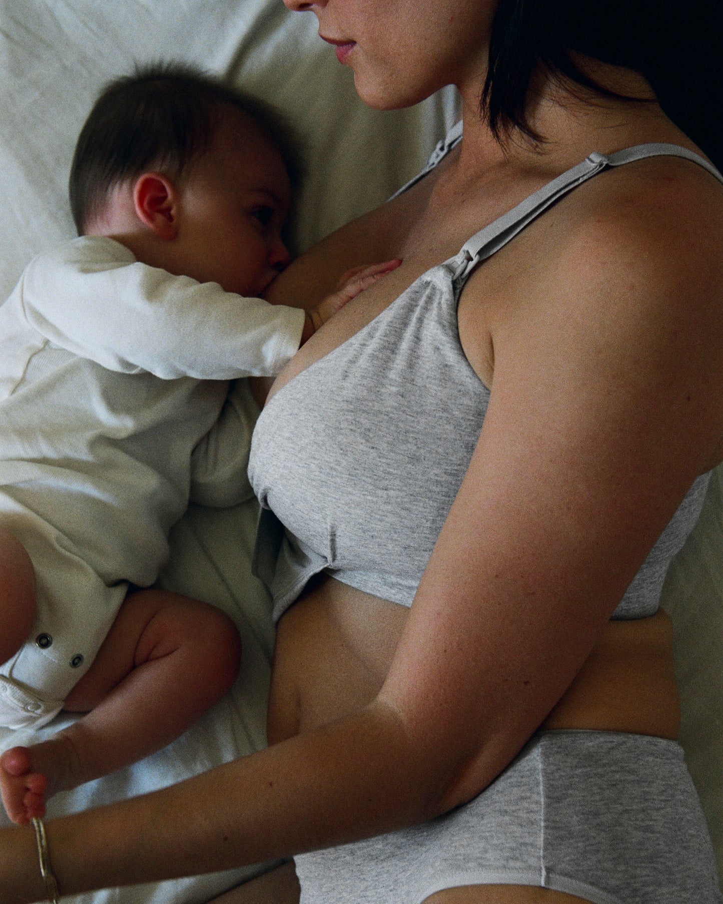 Mother is breastfeeding baby and is wearing SoftStretch Cotton Nursing Bra and SoftStretch Cotton Maternity & Postpartum Hipster in Grey Heather on a white sheet bed in a darkened room.