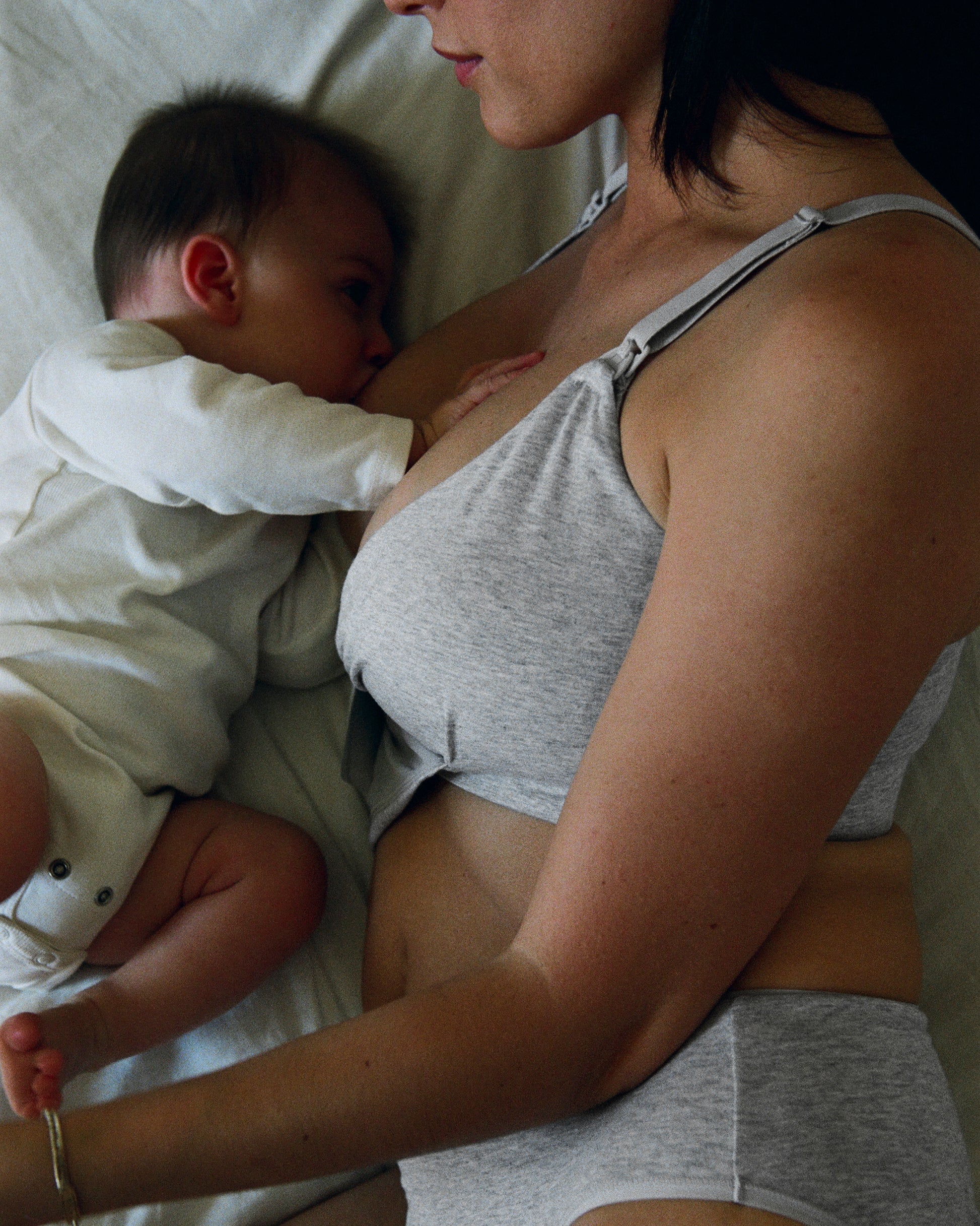 Mother is breastfeeding baby and is wearing SoftStretch Cotton Nursing Bra and SoftStretch Cotton Maternity & Postpartum Hipster in Grey Heather on a white sheet bed in a darkened room.