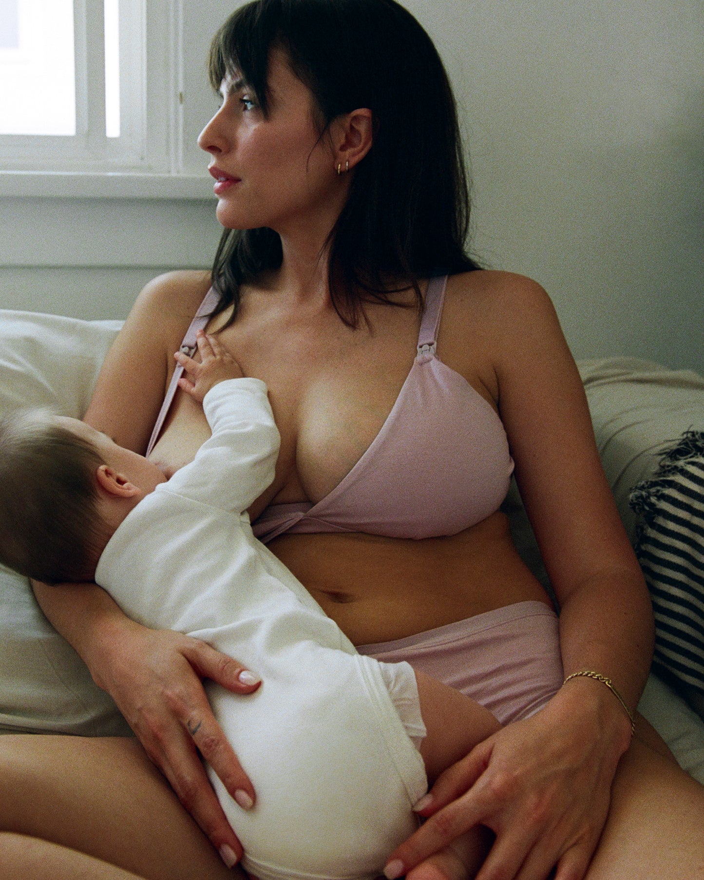 Woman is breastfeeding baby on a chair by the window while wearing SoftStretch Cotton Maternity & Nursing Bralette in Lilac Mist. 