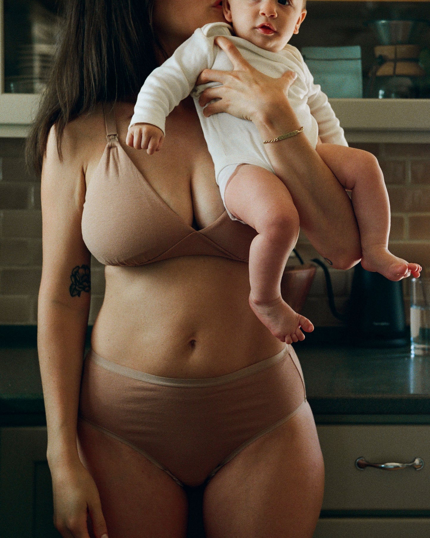 Model is holding and kissing her baby and wearing the SoftStretch Cotton Maternity & Nursing Bralette in Buff, and SoftStretch Cotton Maternity & Postpartum Brief in Buff in a dark kitchen.