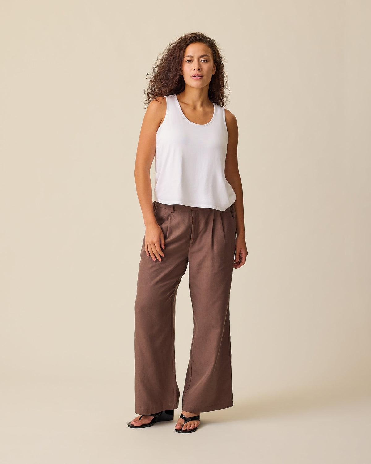 Model is wearing Maternity & Postpartum Wide Leg Trouser in Brown and Ruched Maternity Sleeveless Top in White, on a beige background.