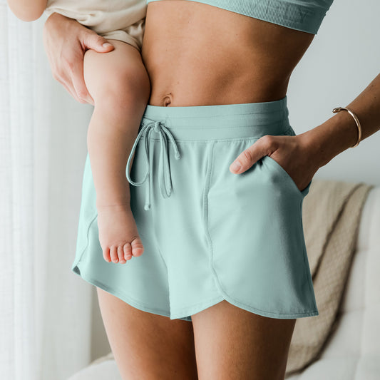3/4 angle view of model wearing the  Bamboo Maternity & Postpartum Lounge Shorts in dusty blue green, holding baby