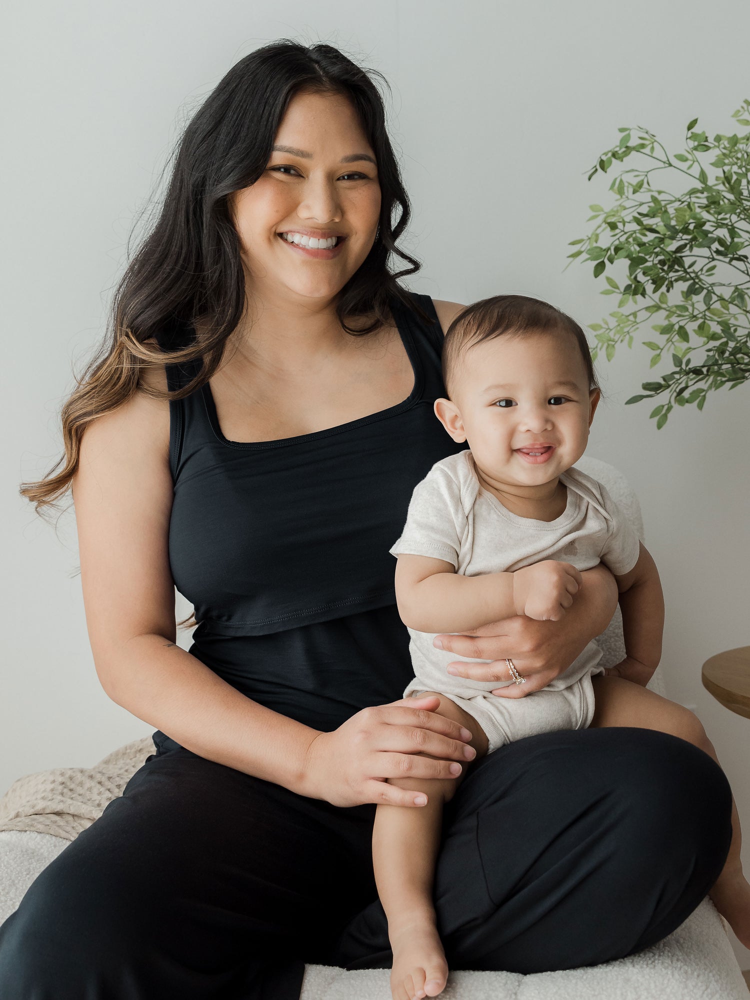 Model with her baby sitting on her lap while wearing the Everyday Essential Nursing Tank in Black. @model_info:Binc is wearing a Medium. 